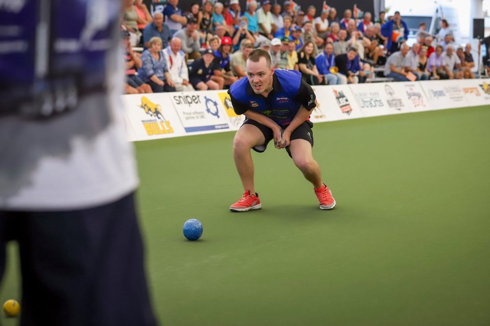 Player crouching to watch his bowl run, with a packed grandstand behind