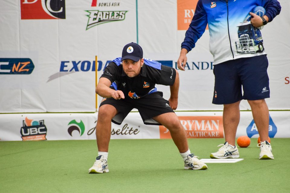 Bowler crouching to watch his delivery as a teammate stands beside the mat