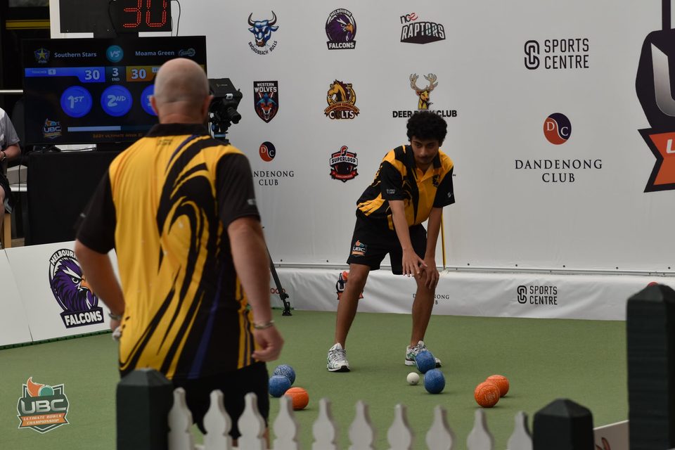 Player in yellow and black stooping to watch his bowl as a teammate looks on