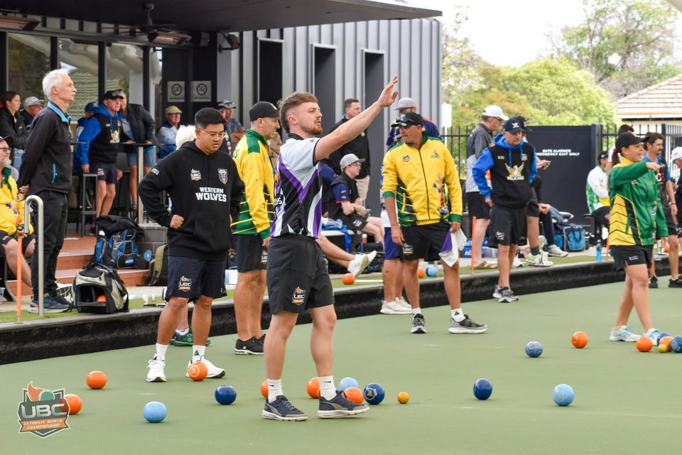Player raising an arm on a busy outdoor green with teams and spectators behind