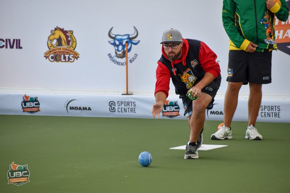 Bowler in a red and black hoodie delivering a bowl in front of the sponsor banners